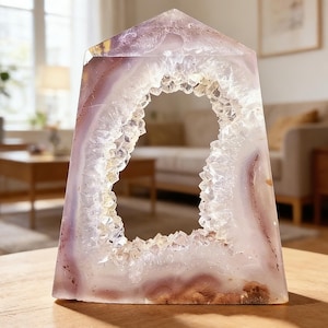 May include: A polished, geode-filled crystal obelisk. The crystal features a hollow center lined with sparkling quartz crystals. The outer layers display bands of lavender, white, and brown hues, creating a natural, decorative piece.