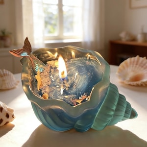 Pode incluir: Uma vela em forma de concha com um detalhe de cauda de sereia. A vela tem um interior de cera azul e transparente com um pavio aceso. A cauda de sereia é castanha e azul-petróleo. Conchas no fundo.