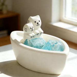 May include: A white ceramic bowl holds blue crystals and two white cat figurines. The cats have black eyes and paws. The bowl is oval-shaped and sits on a white surface.