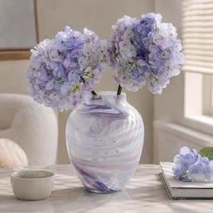 May include: A glass vase with a swirling purple and white marbled pattern containing two large stems of light purple and blue hydrangea flowers. The vase has a rounded body and a narrow neck, displaying a smooth, glossy finish throughout the glass.