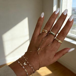 May include: A hand adorned with gold jewelry, including multiple rings and bracelets. The rings feature various designs, such as a band with small stones and a rectangular signet ring. The bracelets include delicate chains with charms and a hand chain.