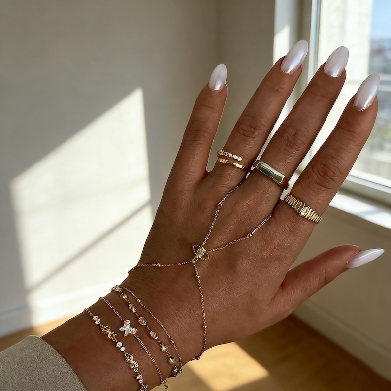May include: A hand adorned with gold jewelry, including multiple rings and bracelets. The rings feature various designs, such as a band with small stones and a rectangular signet ring. The bracelets include delicate chains with charms and a hand chain.
