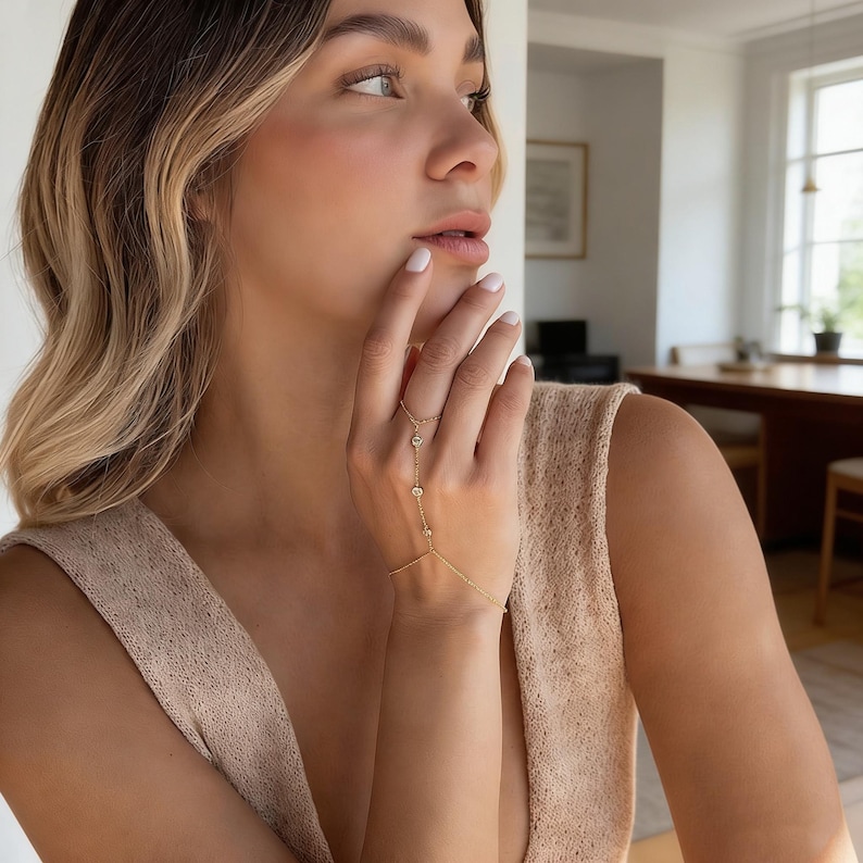 May include: A delicate gold hand chain with three clear, round gemstones. The chain connects to a ring and a bracelet, creating a stylish accessory. The jewelry is worn on a hand, showcasing its elegant design.