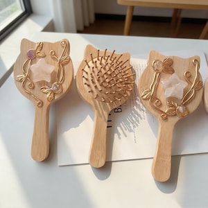 以下が含まれることがあります： 金色の装飾と宝石が付いた、猫の形をした木製のヘアブラシ。1つのブラシは木製の毛、他のブラシは星と宝石の装飾が施されています。ブラシは白い表面に展示されています。