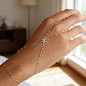 May include: A delicate gold hand chain featuring a floral design. The chain connects to a ring and a bracelet, with a small flower-shaped centerpiece adorned with sparkling stones. The jewelry is designed to be worn on the hand.
