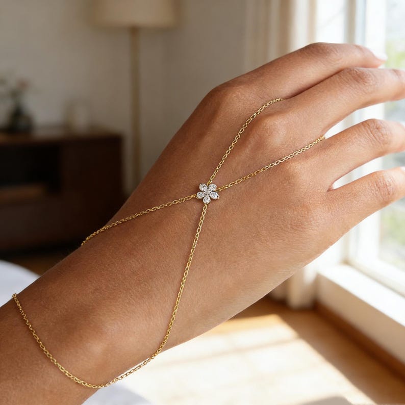 May include: A delicate gold hand chain featuring a floral design. The chain connects to a ring and a bracelet, with a small flower-shaped centerpiece adorned with sparkling stones. The jewelry is designed to be worn on the hand.