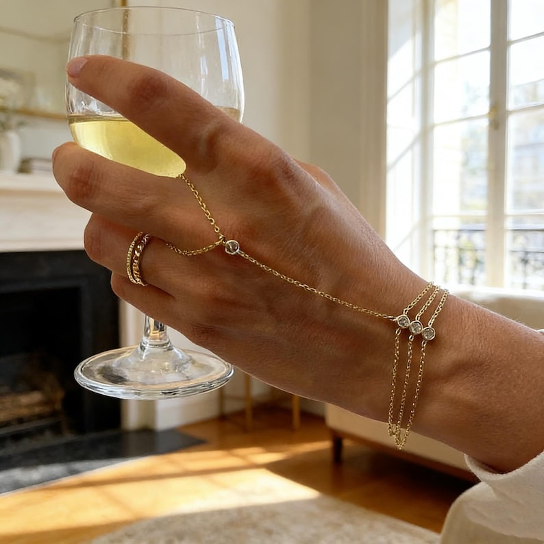 May include: A gold hand chain bracelet with three dangling strands, each adorned with a small, clear gemstone. The chain connects to a ring and a single gemstone on the back of the hand. A glass of white wine is held in the hand.