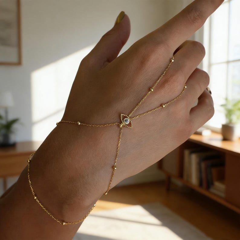 May include: A delicate gold hand chain bracelet. The bracelet features a small, clear gemstone in an eye-shaped setting, connected by fine chains to the wrist and finger. Small gold beads accent the chains. The jewelry is displayed on a hand.