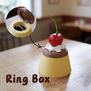 Op de afbeelding: Een gele ringdoos in de vorm van een puddingdessert, bekroond met een rode kers en witte room. De doos is open en onthult een zilveren ketting. De woorden "Ring Box" staan onderaan gedrukt.
