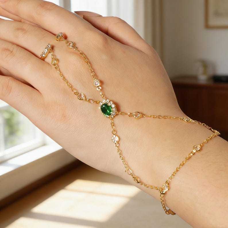 May include: A gold hand chain with a ring and bracelet design. The chain features a central oval green gemstone surrounded by small clear stones. Additional clear stones are set along the chain.