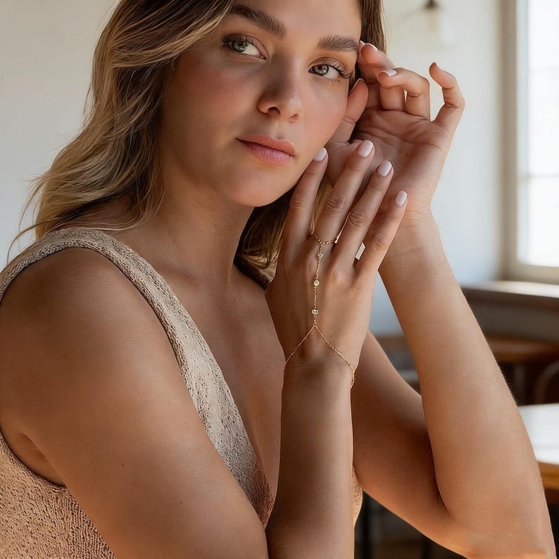 May include: A delicate gold hand chain with multiple small, round, clear gemstones. The chain connects to a ring and wraps around the hand, creating a stylish and elegant look. The jewelry is worn on a hand with manicured nails.