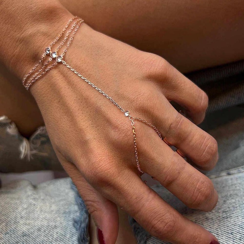 May include: Rose gold hand chain bracelet with three delicate strands on the wrist, connected by a chain to a ring. The bracelet features small, clear gemstones. The jewelry is worn on a hand.