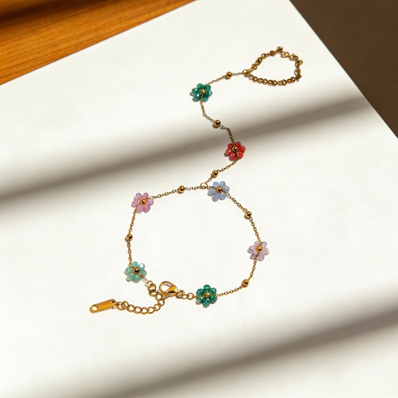 May include: A gold-colored bracelet with a delicate chain design. The bracelet features small, colorful flower-shaped beads in shades of pink, green, red, and blue. The bracelet also has a ring that connects to the bracelet with a chain.