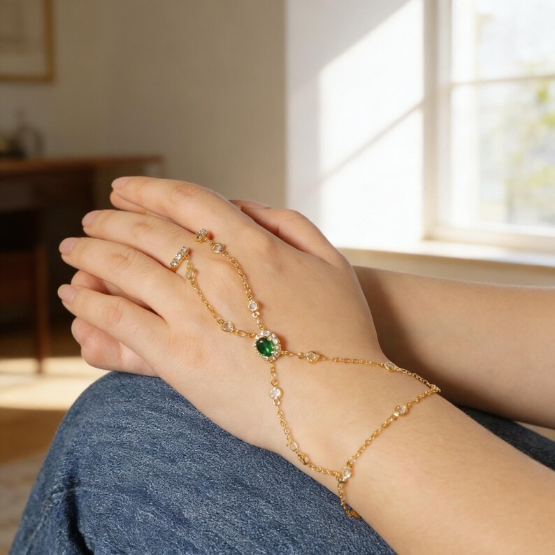May include: A gold-colored hand chain with a ring and a central emerald-green gemstone. The chain is adorned with small, clear gemstones. The jewelry is worn on a hand, with the ring on the index finger.