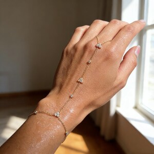 May include: A delicate gold hand chain bracelet with small, sparkling gemstones. The bracelet features a chain that wraps around the wrist and connects to a ring, with decorative stones along the chain. The jewelry is displayed on a hand.