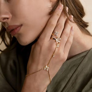 May include: A gold-toned hand chain and ring set. The set features a delicate chain that connects to a ring and a bracelet, adorned with sparkling, clear crystal flower-shaped accents. The ring and bracelet are connected by a fine chain.
