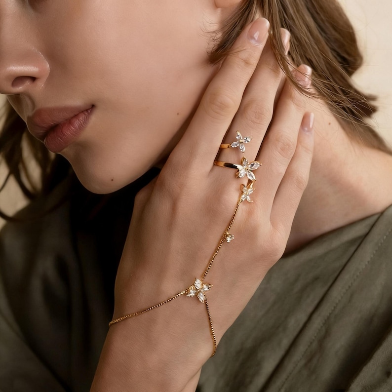 May include: A gold-toned hand chain and ring set. The set features a delicate chain that connects to a ring and a bracelet, adorned with sparkling, clear crystal flower-shaped accents. The ring and bracelet are connected by a fine chain.