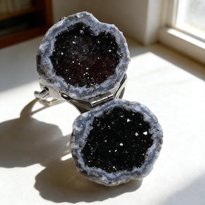May include: A hinged, silver-toned ring featuring two halves of a geode. The geode halves are dark gray with a sparkling interior and a light gray and white border. The ring is open, revealing the geode's interior.