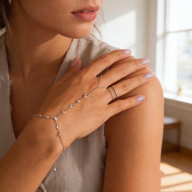 May include: A silver hand chain with a ring and bracelet design. The chain features multiple clear, sparkling gemstones. The ring is a simple band. The jewelry is worn on a person's hand.