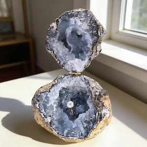 May include: A geode ring box, open to reveal a diamond ring. The box is made of a natural, rough-edged stone with a blue-grey crystal interior. The ring is set in a gold band. The top half of the geode is open, revealing the interior.