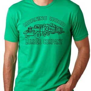 May include: A green t-shirt featuring a black graphic of a vintage logging truck carrying logs. The text "MORNING WOOD LUMBER COMPANY" is printed above and below the truck illustration. The shirt has short sleeves and a crew neck.