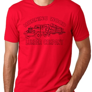 May include: A red t-shirt featuring a black graphic of a vintage logging truck hauling logs. The text "MORNING WOOD LUMBER COMPANY" is arched above and below the truck illustration. The shirt is made of a soft material.