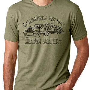 May include: Olive green t-shirt featuring a black graphic of a vintage logging truck carrying logs. The text "MORNING WOOD LUMBER COMPANY" is arched above and below the truck illustration. The shirt is short-sleeved and has a crew neck.