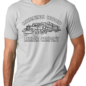 May include: A light gray t-shirt featuring a black graphic of a vintage truck hauling logs. The text "MORNING WOOD LUMBER COMPANY" is arched above and below the truck illustration.
