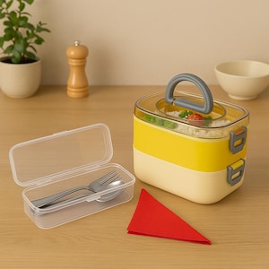May include: A two-tiered yellow and cream-colored bento box with a gray handle, filled with food. A clear plastic container holds a fork and spoon. A red napkin sits nearby. A small plant and a bowl are in the background.