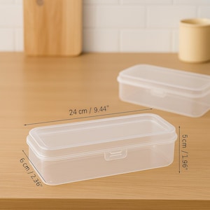 May include: Clear rectangular plastic storage container with a lid. The container is 9.44 inches long, 2.36 inches wide, and 1.96 inches tall. The container is empty and sits on a light brown surface.