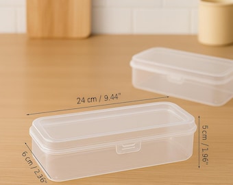 Può includere: Contenitore di stoccaggio rettangolare in plastica trasparente con coperchio. Il contenitore è lungo 24 cm, largo 6 cm e alto 5 cm. Il contenitore è vuoto e si trova su una superficie marrone chiaro.