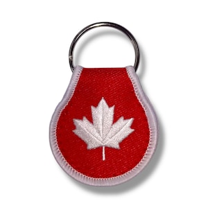 May include: A red fabric keychain with a white embroidered maple leaf. The keychain has a silver metal ring.