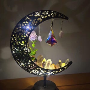 May include: A black crescent moon-shaped lamp with intricate cutouts, illuminated from within. The lamp features a central iridescent geometric crystal pendant, a small artificial flower, and a collection of clear quartz-like crystals at the base.