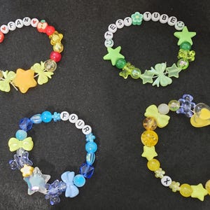 May include: Four colorful beaded bracelets with different charms and letter beads spelling out "FAN", "TEST TUBE", "FOUR", and a heart with an "X" on it.