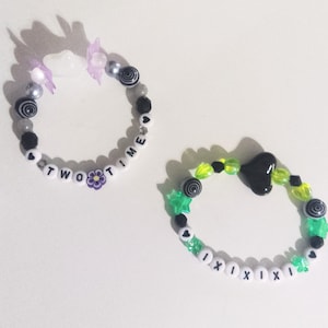 May include: Two beaded bracelets. One bracelet has the words "TWO TIME" and a purple flower charm. The other bracelet has the word "XIXIXI" and a black heart charm. Both bracelets have a variety of beads in black, white, and other colors.