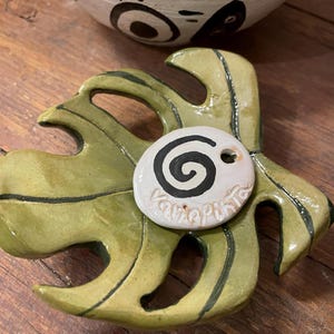 Può includere: Un piatto in ceramica verde a forma di foglia con un'etichetta in ceramica bianca che dice "Varepunta" e ha un design a spirale nero.