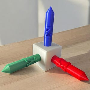 May include: A 3D-printed geometric sculpture featuring a white cube with three colored, pointed cylinders. One cylinder is blue and upright, while the other two are red and green, extending outward. The sculpture has a modern, abstract design.