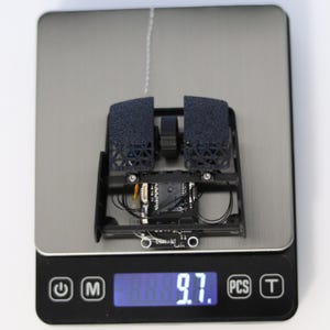 May include: A black and gray 3D printed mouse with a circuit board and wires visible. The mouse is on a digital scale that reads 9.7.