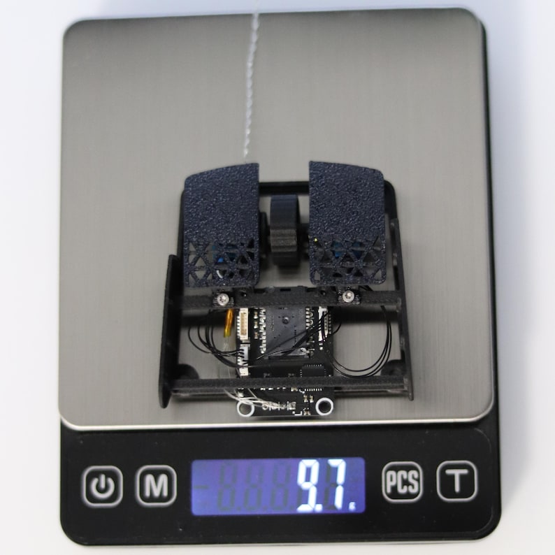 May include: A black and gray 3D printed mouse with a circuit board and wires visible. The mouse is on a digital scale that reads 9.7.