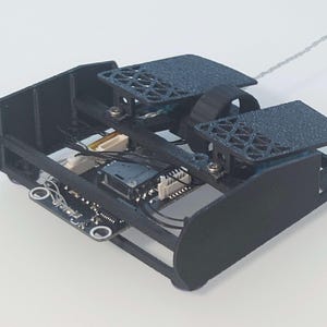 May include: A black 3D printed robot with a circuit board and wires visible. The robot has two wheels and a small camera.