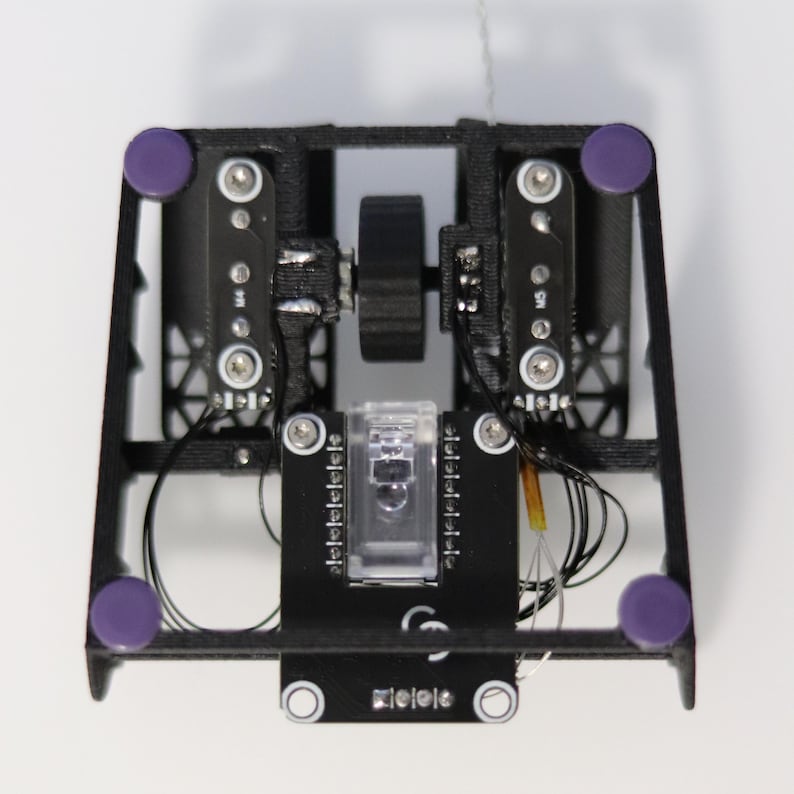 May include: A black 3D printed device with a clear plastic box, a small motor, and two purple buttons. The device has a circuit board with wires and a small orange wire.