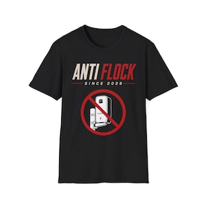 May include: A black short-sleeved t-shirt featuring the text ANTI FLOCK SINCE 2026 in white and red lettering. Below the text is a graphic of a surveillance camera mounted on a pole, enclosed within a red circular prohibition symbol with a diagonal line.