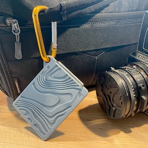 May include: A light blue rectangular tag featuring a raised topographic map pattern on its surface. The tag is attached to a gold-colored metal carabiner clip, which is hooked onto the strap of a black camera bag next to a black camera lens.