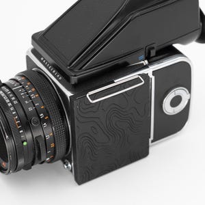 May include: A close-up of a Hasselblad medium format camera. The camera is primarily black with silver accents and a textured black panel. The lens has various measurement markings. The Hasselblad logo is visible.