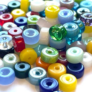 May include: A collection of small, round, disc-shaped glass beads in a variety of solid and translucent colors including red, yellow, blue, green, white, and purple. Each bead features a single centered hole, creating a uniform, flat, circular profile.