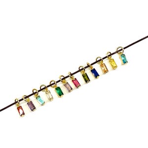 May include: A collection of gold-colored charms, each featuring a rectangular, multi-colored gemstone. The charms are suspended from a thin, dark wire, showcasing a spectrum of colors including red, purple, green, and blue. The background is plain white.