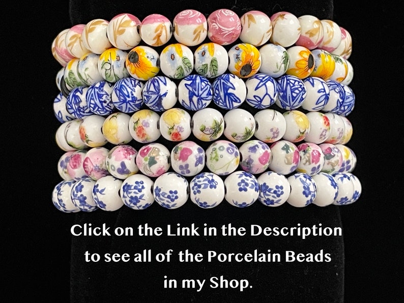 Pu&ograve; includere: Una collezione di perline rotonde in porcellana con vari disegni floreali e geometrici. Le perline sono bianche con dettagli dipinti a mano in rosa, giallo, blu e verde. L'immagine include il testo: "Click on the Link in the Description to see all of the Porcelain Beads in my Shop."
