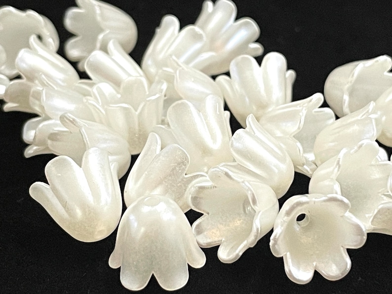 May include: A group of white plastic flower beads with a pearlescent finish. The beads are shaped like a bell with five petals.