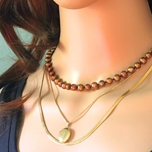 May include: A layered necklace set featuring a gold chain with a locket pendant, a thinner gold chain, and a strand of brown pearls.