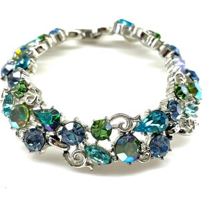 May include: A silver-toned metal bracelet featuring a series of multi-shaped faceted crystals in shades of blue, teal, and green. The crystals are set within ornate, swirling metal links that connect to form a circular, flexible band design.